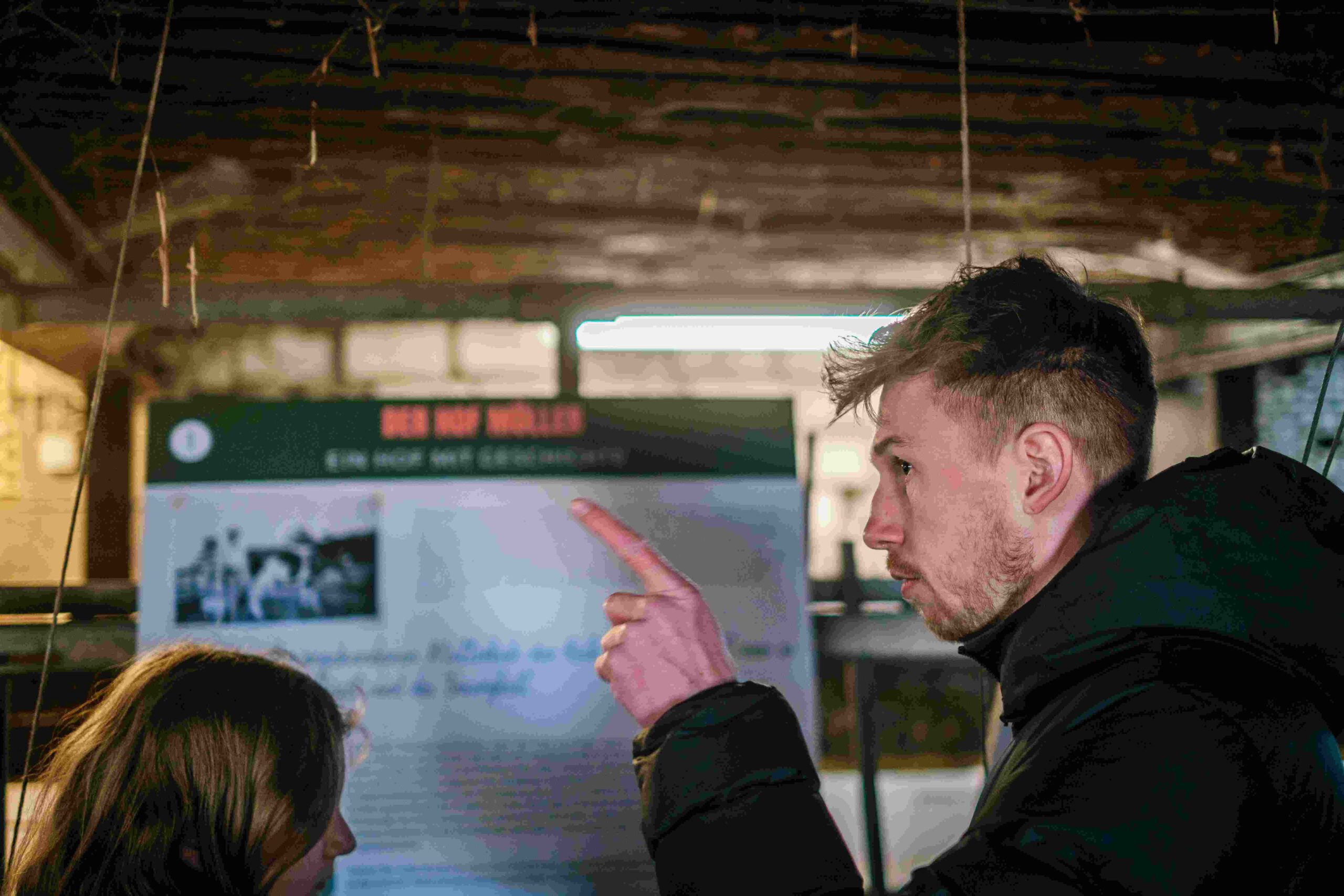 Ein Mann in dunkler Kleidung steht in einer Scheune oder Halle und zeigt mit seinem Finger auf eine Infotafel im Hintergrund. Neben ihm ist eine weitere Person von hinten zu sehen. Die Tafel trägt den Titel „Der Hof Möller“ und zeigt Bilder und Text. Die Umgebung ist schummrig beleuchtet, mit Stroh oder Seilen, die von der Decke hängen.
