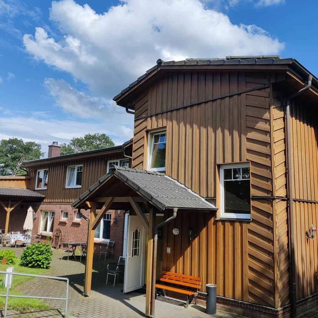Der Ferienhof Möllers Morgen mit einer Holzfassade und einem kleinen überdachten Eingangsbereich. Neben der Eingangstür steht eine Holzbank, und links ist ein gepflasterter Hof mit Tischen, Stühlen und einem Sonnenschirm zu sehen. Im Hintergrund verbindet sich die Holzfassade mit einer roten Klinkerwand. Der Himmel ist blau mit weißen Wolken.