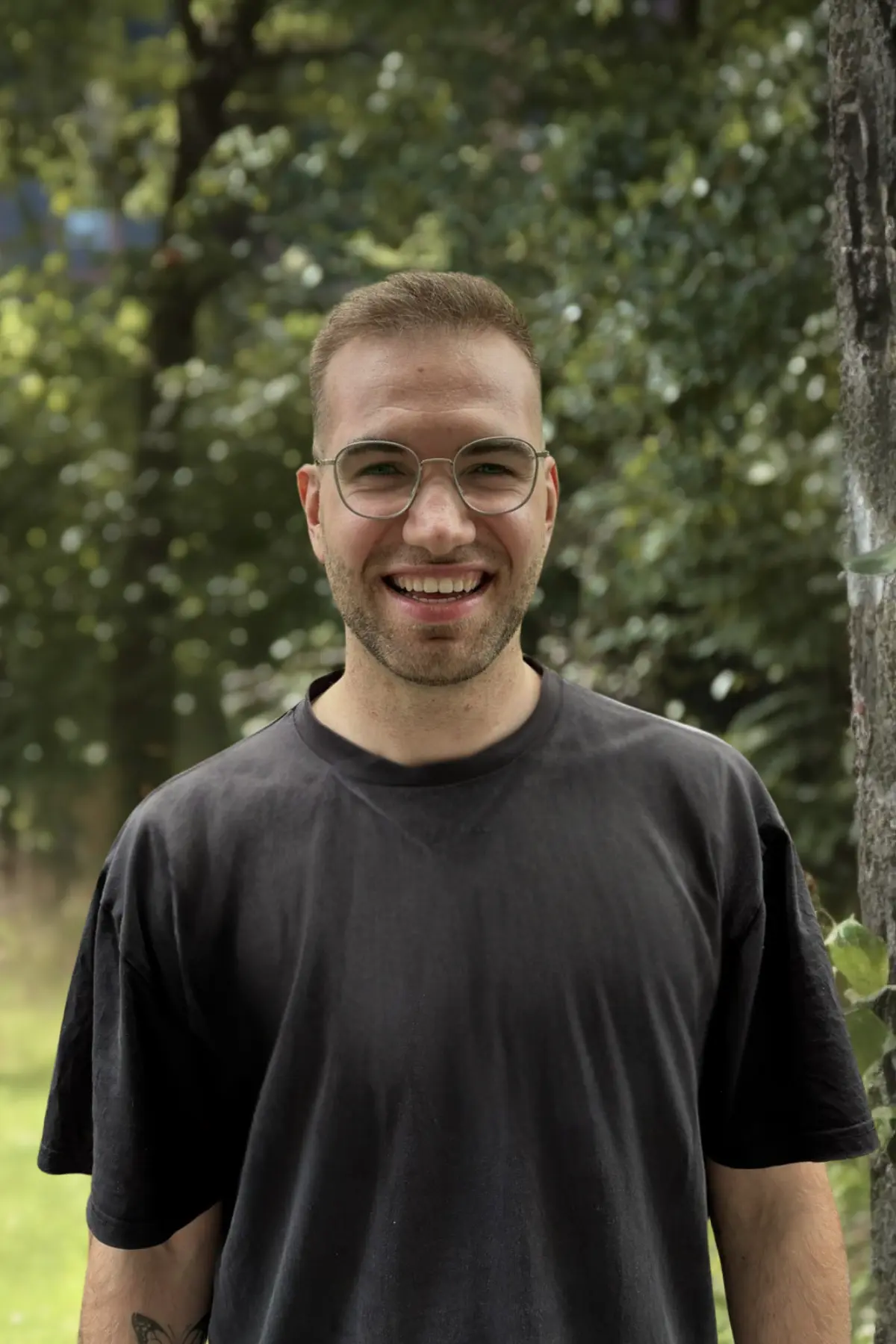 Person trägt ein schwarzes T-Shirt und eine Brille mit dünnem Rahmen. Sie lächelt vor einem unscharfen Hintergrund aus grünen Bäumen.