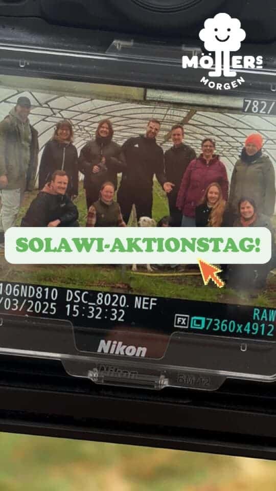 SOLAWI-AKTIONSTAG! Eine Gruppe von Menschen posiert lächelnd vor einem Gewächshaus, fotografiert mit einer Nikon-Kamera. Das Bild zeigt die Kameraeinstellungen und das Datum 03/2025.