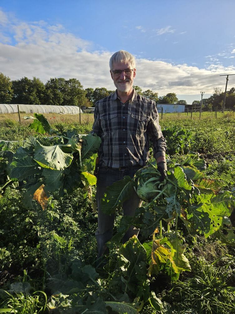 Ein Mann steht in einem Garten zwischen großen grünen Pflanzen, die wie Kürbisse aussehen. Im Hintergrund sind ein Gewächshaus und ein offenes Feld zu sehen.