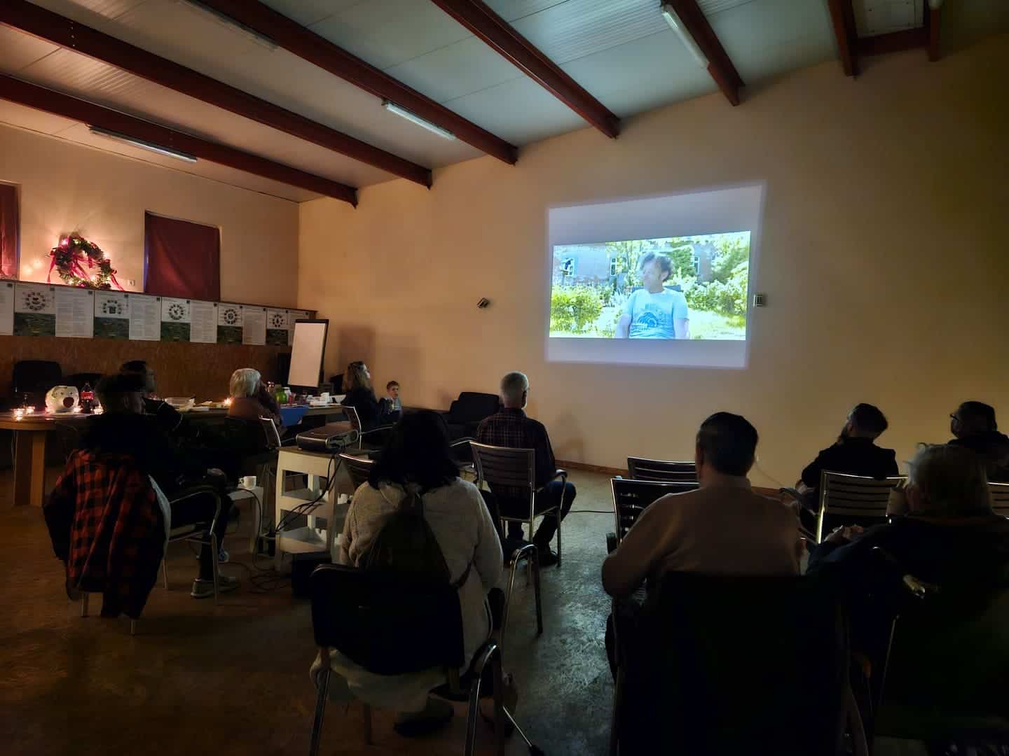 Ein Raum, in dem eine Präsentation oder ein Vortrag stattfindet. Mehrere Personen sitzen auf Stühlen und schauen auf eine Leinwand, auf der ein Bild einer Person zu sehen ist, die in einem Garten oder einer natürlichen Umgebung steht. Der Raum hat eine warme Beleuchtung und eine gemütliche Atmosphäre, mit Dekorationen an den Wänden.