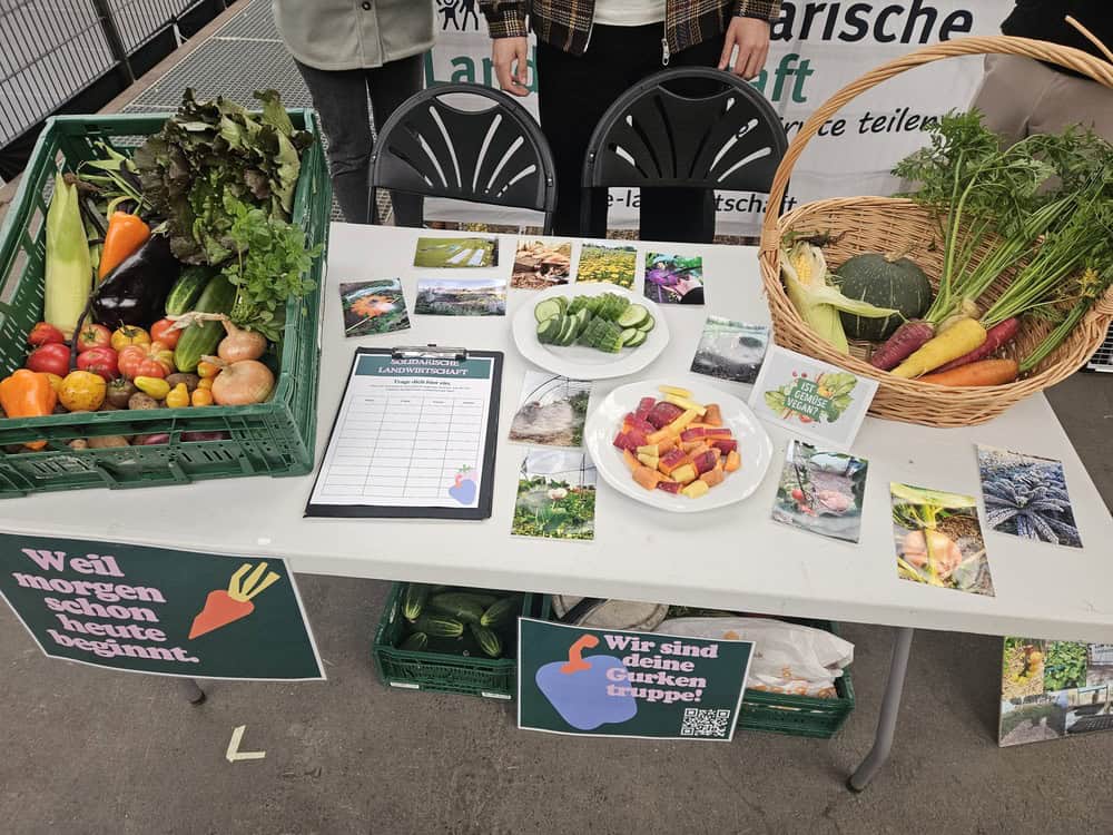 Ein Tisch mit verschiedenen Gemüsesorten, darunter Tomaten, Gurken, Karotten und Kürbisse, sowie Informationsmaterialien und Fotos. Auf dem Tisch befinden sich auch ein Klemmbrett mit einem Formular und Schilder mit Aufschriften wie "Weil morgen schon heute beginnt" und "Wir sind deine Gurken-Truppe!" Zwei Personen stehen hinter dem Tisch.
