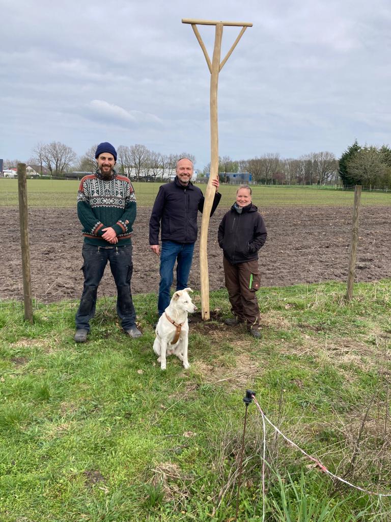 Drei Männer und ein Hund stehen auf einem Feld, wobei einer der Männer einen hölzernen Pfosten hält. Im Hintergrund sind Bäume und ein offenes Feld zu sehen.