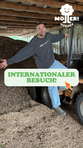 Das Bild zeigt eine Person, die auf einem Gabelstapler sitzt und sich in einer Lagerhalle oder einem landwirtschaftlichen Betrieb befindet. Die Person trägt ein graues Sweatshirt und Jeans und lächelt in die Kamera. Im Hintergrund ist ein großer Haufen von Holzspänen oder Mulch zu sehen, was darauf hindeutet, dass es sich um einen Betrieb handelt, der mit Holz oder organischen Materialien arbeitet. Über dem Bild ist ein Text mit der Aufschrift "Internationaler Besuch!" zu sehen, was darauf hindeutet, dass die Person möglicherweise internationale Gäste empfängt oder auf einen internationalen Besuch hinweist. Das Logo "Möllers Morgen" ist in der oberen rechten Ecke des Bildes sichtbar, was darauf hinweist, dass die Person möglicherweise mit diesem Betrieb verbunden ist.