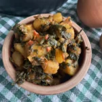 Eine Schüssel mit einer leckeren Kartoffel-Mangold-Pfanne. Das Gericht besteht aus goldbraunen Kartoffelstücken und frischem, grünem Mangold. Die Pfanne ist in einer rustikalen Keramikschüssel angerichtet und wirkt frisch zubereitet. Im Hintergrund sind eine Pfanne und eine Zwiebel auf einem karierten Küchentuch zu sehen.