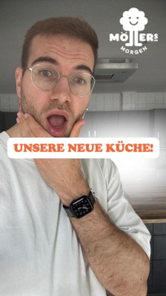 Das Bild zeigt eine Person, die in einer Küche steht und überrascht oder aufgeregt wirkt. Die Person trägt ein weißes T-Shirt und eine Brille und hat eine Hand an ihr Gesicht gelegt, während sie in die Kamera schaut. Sie trägt eine Smartwatch am Handgelenk. Im Hintergrund ist eine moderne Küche mit weißen Schränken und einer Dunstabzugshaube zu sehen. Über dem Bild ist ein Text mit der Aufschrift "Unsere neue Küche!" zu sehen, was darauf hindeutet, dass die Küche neu eingerichtet oder renoviert wurde. Das Logo "Möllers Morgen" ist in der oberen rechten Ecke des Bildes sichtbar, was darauf hinweist, dass die Person möglicherweise mit diesem Betrieb verbunden ist.