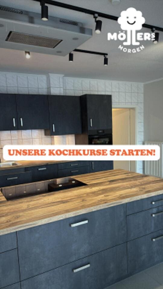 Das Bild zeigt eine moderne Küche mit einer großen Insel in der Mitte. Die Küchenschränke sind in einem dunklen Grauton gehalten, während die Arbeitsplatte der Insel aus hellem Holz besteht. Die Küche ist mit modernen Geräten ausgestattet, darunter ein Kochfeld auf der Insel und eine Dunstabzugshaube darüber. Die Beleuchtung besteht aus mehreren kleinen, modernen Deckenleuchten, die an einer Schiene befestigt sind. Über dem Bild ist ein Text mit der Aufschrift "Unsere Kochkurse starten!" zu sehen, was darauf hindeutet, dass Kochkurse in dieser Küche angeboten werden. Das Logo "Möllers Morgen" ist in der oberen rechten Ecke des Bildes sichtbar, was darauf hinweist, dass die Küche mit diesem Betrieb verbunden ist.