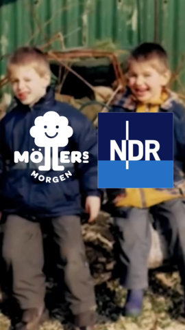 Das Bild zeigt zwei Kinder, die draußen auf einem Haufen von Holzspänen oder Mulch sitzen und in die Kamera lächeln. Die Kinder tragen wetterfeste Kleidung, darunter eine blaue Jacke mit dem Logo "Möllers Morgen" und eine gelbe Jacke. Im Hintergrund ist ein grünes Wellblech zu sehen, das möglicherweise zu einem Gebäude oder einer Scheune gehört. Das Bild vermittelt eine fröhliche und spielerische Atmosphäre, während die Kinder draußen spielen. Das Logo des NDR ist ebenfalls im Bild zu sehen, was darauf hinweist, dass das Bild möglicherweise im Zusammenhang mit einer Sendung oder einem Bericht des NDR steht.