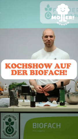 Das Bild zeigt eine Person, die eine Kochshow auf der BioFach präsentiert, einer internationalen Messe für Bio-Lebensmittel. Die Person steht hinter einem Tisch, auf dem verschiedene Zutaten und Küchenutensilien wie Flaschen mit Öl, Kräuter, Schüsseln und Schneidebretter zu sehen sind. Sie scheint aktiv am Kochen oder Vorbereiten eines Gerichts beteiligt zu sein und hält ein Mikrofon in der Hand, was darauf hindeutet, dass sie gleichzeitig spricht oder Anweisungen gibt. Über dem Bild ist ein Text mit der Aufschrift "Kochshow auf der Biofach!" zu sehen, was den Kontext der Kochshow auf der BioFach-Messe verdeutlicht. Im Hintergrund ist ein Banner mit dem Logo "Möllers Morgen" sichtbar, was darauf hinweist, dass die Person oder die Kochshow mit diesem Betrieb verbunden ist.
