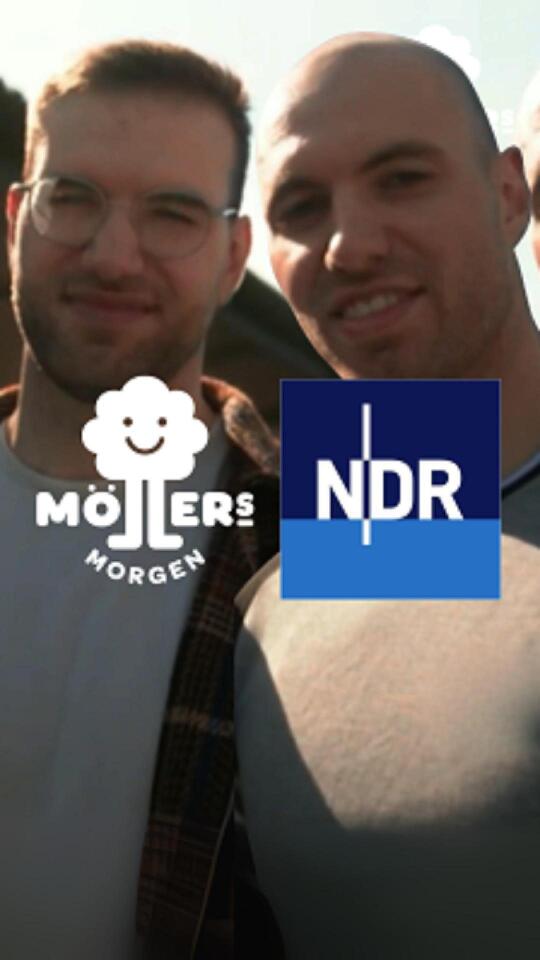 Das Bild zeigt zwei Personen, die sich für ein Selfie zusammen posieren. Die Person auf der linken Seite trägt eine Brille und ein hellgraues T-Shirt mit dem Logo "Möllers Morgen", während die Person auf der rechten Seite ein dunkles T-Shirt trägt. Beide Personen lächeln in die Kamera. Im Vordergrund des Bildes ist das Logo des NDR zu sehen, was darauf hinweist, dass das Bild möglicherweise im Zusammenhang mit einer Sendung oder einem Bericht des NDR steht.