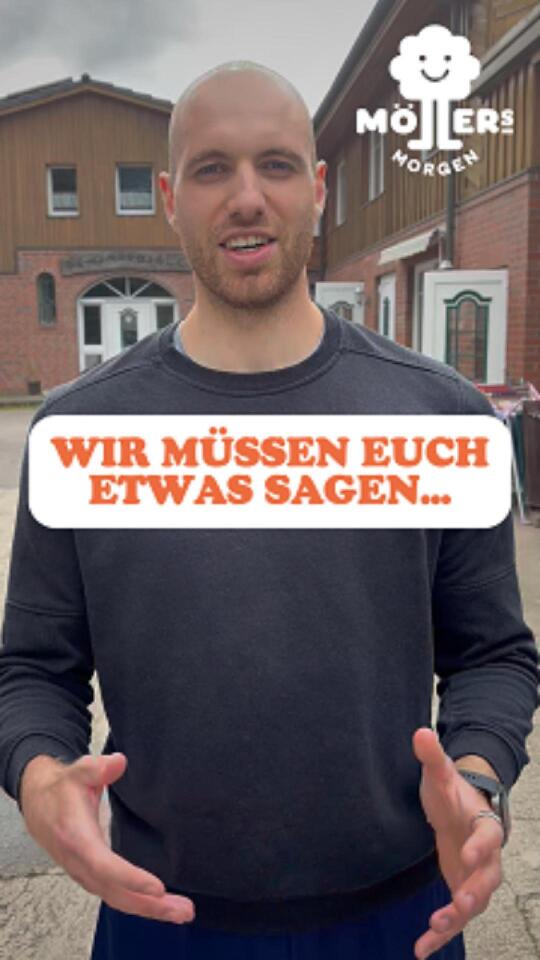 Das Bild zeigt eine Person, die vor einem Gebäude steht und in die Kamera spricht. Die Person trägt ein dunkles Sweatshirt und hat eine freundliche, offene Körpersprache mit ausgestreckten Händen. Über dem Bild ist ein Text mit der Aufschrift "Wir müssen euch etwas sagen..." zu sehen, was darauf hindeutet, dass die Person eine wichtige Mitteilung oder Ankündigung machen möchte. Im Hintergrund ist ein Gebäude mit einer traditionellen Fassade zu sehen, und in der oberen rechten Ecke des Bildes ist das Logo "Möllers Morgen" sichtbar, was darauf hinweist, dass die Person mit diesem Betrieb verbunden ist.