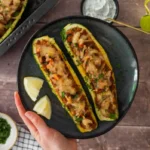 Ein dunkler Teller mit zwei halbierten und gefüllten Zucchiniboaten, die mit veganem Hackfleisch, Tomaten, Zwiebeln und Kräutern gefüllt sind. Das Gericht ist überbacken und mit frischen Kräutern bestreut. Dazu gibt es zwei Zitronenspalten und eine kleine Schale mit Joghurt-Dip. Im Hintergrund liegt eine Backform mit weiteren gefüllten Zucchini, und goldene Essbesteckteile sind auf einem Holztisch arrangiert.