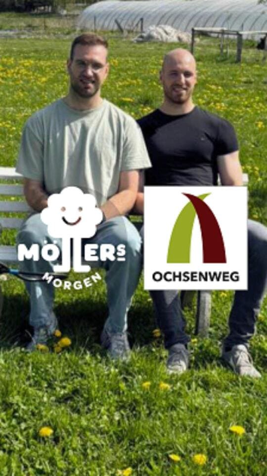 Zwei lächelnde Männer sitzen auf einer weißen Bank vor einem Feld mit blühenden Pflanzen und Gewächshäusern im Hintergrund. Der Mann auf der linken Seite trägt ein hellgraues T-Shirt, der Mann auf der rechten Seite ein schwarzes T-Shirt. Im Bild sind die Logos von Möllers Morgen und Ochsensenweg eingeblendet. Die Szene wirkt freundlich und ländlich.