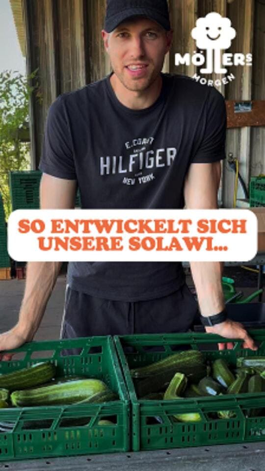Ein lächelnder Mann mit Basecap und einem schwarzen T-Shirt mit der Aufschrift "HILFIGER" steht vor einer Kiste mit frischen Zucchini. Im Hintergrund ist ein Gewächshaus oder ein Hofladen zu sehen. Über dem Bild steht in großen, orangefarbenen Buchstaben: SO ENTWICKELT SICH UNSERE SOLAWI... Das Logo von Möllers Morgen ist in der oberen rechten Ecke sichtbar.