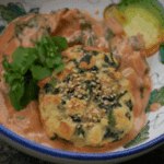 Ein Teller mit Spinatknödeln, die mit einer cremigen, orangefarbenen Sauce übergossen sind. Die Knödel sind mit Sesam- oder Sonnenblumenkernen bestreut und werden von frischem Salat oder Kräutern garniert. Dazu gibt es eine Scheibe gebratener Zucchine.