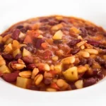 Eine weiße Schüssel mit bunten, dickflüssigen Bohneneintopf, der Kidneybohnen, weiße Bohnen, Kartoffelstücke, Karotten und Tomaten enthält. Der Eintopf wirkt herzhaft und farbenfroh.
