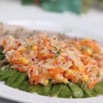 Ein ovaler, weißer Teller mit buntem Zuckerschotensalat, der frische Zuckerschoten, geraspelte Möhren, Maiskörner und eine würzige Marinade enthält. Der Salat ist mit Kräutern bestreut und wird von einem Stück Brot begleitet. Das Gericht wirkt frisch und knackig.