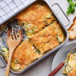 Eine Metall-Bratpfanne mit schnellem veganem Zucchini-Gratin, das eine goldbraune Knusperkruste hat. Das Gratin ist in Stücke geschnitten und wird mit einem Holzlöffel serviert. Daneben liegt ein Teller mit einer Portion des Gratins, Gabel und Messer, sowie zwei Stücke geröstetes Brot. Ein paar frische Basilikumblätter sind zur Dekoration danebengelegt.