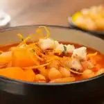 Eine schwarze Schüssel mit herzhaftem Kartoffel-Möhren-Kichererbsen-Eintopf, garniert mit dünnen Orangenzesten und kleinen Stückchen von Fischersatz oder Tofu. Der Eintopf wirkt würzig und farbenfroh.