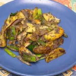 Ein blauer Teller mit gebratenen Kartoffelstücken in einer würzigen Currymarinade, kombiniert mit grünen Paprikastreifen und Frühlingszwiebeln, bestreut mit Mohnsamen. Das Gericht wirkt aromatisch und knusprig.