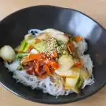 Eine schwarze Schüssel mit Glasnudelsalat, der Pak Choi, Karotten, Gurkenstreifen und eine würzige Sauce enthält. Das Gericht ist mit Sesamsamen bestreut und farbenfroh angerichtet.
