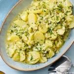 Ein ovaler, blauer Teller mit Uschis Kartoffel-Gurkensalat, bestehend aus gewürfelten, gekochten Kartoffeln, Gurkenstücken und einer cremigen, grünen Kräutermarinade. Der Salat ist frisch angerichtet und wird von zwei Löffeln begleitet. Das Gericht wirkt erfrischend und herzhaft.