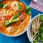 Das Bild zeigt eine Schüssel mit veganer Thai-Curry-Glasnudelsuppe, gefüllt mit Glasnudeln, Gurkenstreifen, roten Zwiebeln, Paprika und Frühlingszwiebeln in einer würzigen, orangefarbenen Currybrühe. Daneben befindet sich eine Schale mit frischen Sojasprossen und frischem Koriandergrün. Das Gericht ist ansprechend angerichtet und wird auf einem blauen Holztisch serviert.