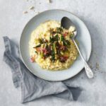 Das Bild zeigt einen Teller mit **Risotto, das mit Catalogna, Pinienkernen und Rosinen** verfeinert ist. Das Risotto ist cremig und gelb, während der Catalogna – eine leicht bittere Chicorée-Art – in Streifen geschnitten und mit gerösteten Pinienkernen und Rosinen kombiniert ist. Die Kombination aus süßen Rosinen, knusprigen Pinienkernen und dem herzhaften Catalogna verleiht dem Gericht eine ausgewogene und aromatische Note. Das Gericht wirkt ansprechend und perfekt für ein raffiniertes, vegetarisches Essen.