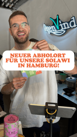 Ein lächelnder Mann mit Brille hält eine bunte Karte in der Hand und steht vor einem Handy-Stativ, das für eine Aufnahme vorbereitet ist. Im Hintergrund hängt ein Neon-Schild mit der Aufschrift "Gegen Wind". Im Vordergrund steht in großen, orangefarbenen Buchstaben: **"NEUER ABHOLORT FÜR UNSERE SOLAWI IN HAMBURG!"** Die Szene wirkt freundlich und informativ, als würde er eine gute Nachricht über den neuen Abholort für die Solidarische Landwirtschaft (Solawi) in Hamburg verkünden.