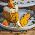 Das Bild zeigt ein Stück veganen Rüblikuchen auf einem Teller, garniert mit pflanzlicher Frischkäseglasur und kleinen Karotten als Dekoration. Der Kuchen sieht saftig und appetitlich aus. Falls gewünscht, kann ich dir ein Rezept dafür bereitstellen.