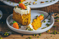 Das Bild zeigt ein Stück veganen Rüblikuchen auf einem Teller, garniert mit pflanzlicher Frischkäseglasur und kleinen Karotten als Dekoration. Der Kuchen sieht saftig und appetitlich aus. Falls gewünscht, kann ich dir ein Rezept dafür bereitstellen.