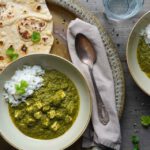Zwei Schalen mit **Palak Paneer mit Tofu** – einem grünen, cremigen Spinat-Curry mit Tofuwürfeln – serviert auf einer dekorativen Metallplatte. Dazu gibt es weiß gekochten Reis und frisches Fladenbrot. Um die Schalen herum sind frische Zutaten wie Knoblauch, Ingwer, Korianderblätter und eine rote Chili zu sehen. Die Szene wirkt aromatisch, farbenfroh und einladend.