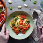Eine Person hält einen Teller mit **Spaghetti al Pomodoro**, garniert mit frischen Basilikumblättern und getrockneten Kirschtomaten. Daneben liegen frische Zutaten wie Karotten, Kirschtomaten, Knoblauchzehen und Basilikumblätter auf einem dunklen Holztisch. Ein Glas Wasser und Besteck sind ebenfalls zu sehen. Das Gericht wirkt frisch, aromatisch und einladend – perfekt für ein gesundes und leckeres Essen!