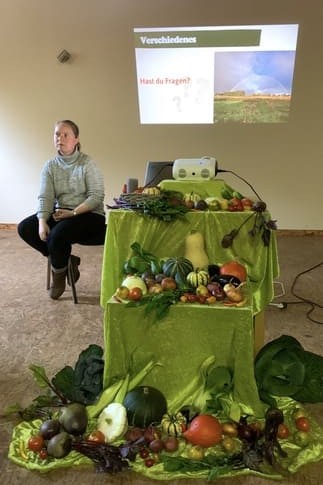 Das Bild zeigt eine Frau, die vor einem Tisch mit einer Vielfalt an frischem, saisonalem Gemüse sitzt. Auf dem Tisch liegt ein grünes Tuch, auf dem verschiedene Sorten wie Kürbisse, Tomaten, Äpfel, Trauben, Rote Bete und Kohlköpfe dekorativ arrangiert sind. Im Hintergrund ist eine Leinwand mit der Frage:
**"Verschwindenes – Hast du Fragen?"**
Die Szene wirkt wie eine Informationsveranstaltung oder ein Workshop zum Thema nachhaltige Landwirtschaft, Gemüseanbau oder Ernährung. Die Frau scheint bereit zu sein, Fragen zu beantworten oder über die Bedeutung von saisonalem und regionalem Gemüse zu informieren.