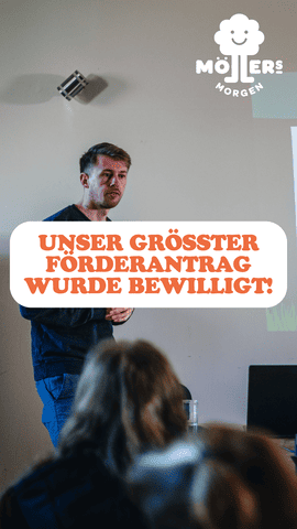 Das Bild zeigt einen Mann, der vor einer Gruppe von Menschen steht und einen Vortrag hält. Im Vordergrund steht in großen, orangefarbenen Buchstaben: **"UNSER GRÖSSTER FÖRDERANTrag WURDE BEWILLIGT!"** Das Logo von **Möllers Morgen** ist in der oberen rechten Ecke sichtbar. Die Szene wirkt wie eine wichtige Ankündigung oder ein Meilenstein für das Projekt oder die Organisation. Der Mann scheint engagiert und freudig die gute Nachricht mit den Anwesenden zu teilen.