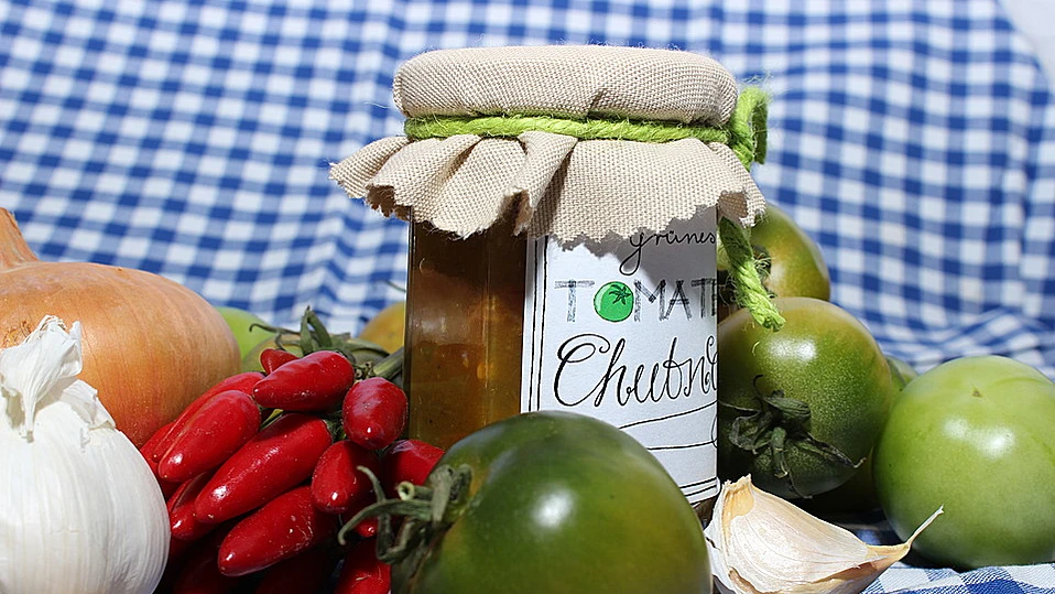 Das Bild zeigt ein Glas mit **veganem Chutney aus grünen Tomaten**, das mit einem Stoffdeckel verschlossen ist. Das Etikett auf dem Glas ist handschriftlich beschriftet und trägt die Aufschrift **"grüne Tomaten Chutney"**. Um das Glas herum liegen frische Zutaten wie grüne Tomaten, Knoblauch, Zwiebeln und rote Chili-Schoten, die typischerweise für die Zubereitung eines Chutneys verwendet werden. Die Szene vermittelt den Eindruck von hausgemachter, nachhaltiger Lebensmittelverarbeitung und lädt zum Probieren ein. Ein perfektes Beispiel für kreative Verwertung von saisonalem Gemüse!