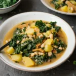 Das Bild zeigt einen Teller mit einem herzhaften **veganen Grünkohleintopf**, der mit Kartoffeln, weißen Bohnen, Pilzen und Grünkohl zubereitet ist. Der Eintopf wirkt cremig und nährstoffreich, garniert mit frischen Kräutern und serviert in einer ansprechenden Schale. Im Hintergrund sind weitere Schüsseln mit Grünkohl zu sehen, was die frische und saisonale Zutatenwahl unterstreicht. Ein perfektes Gericht für eine gesunde, vegane Ernährung!