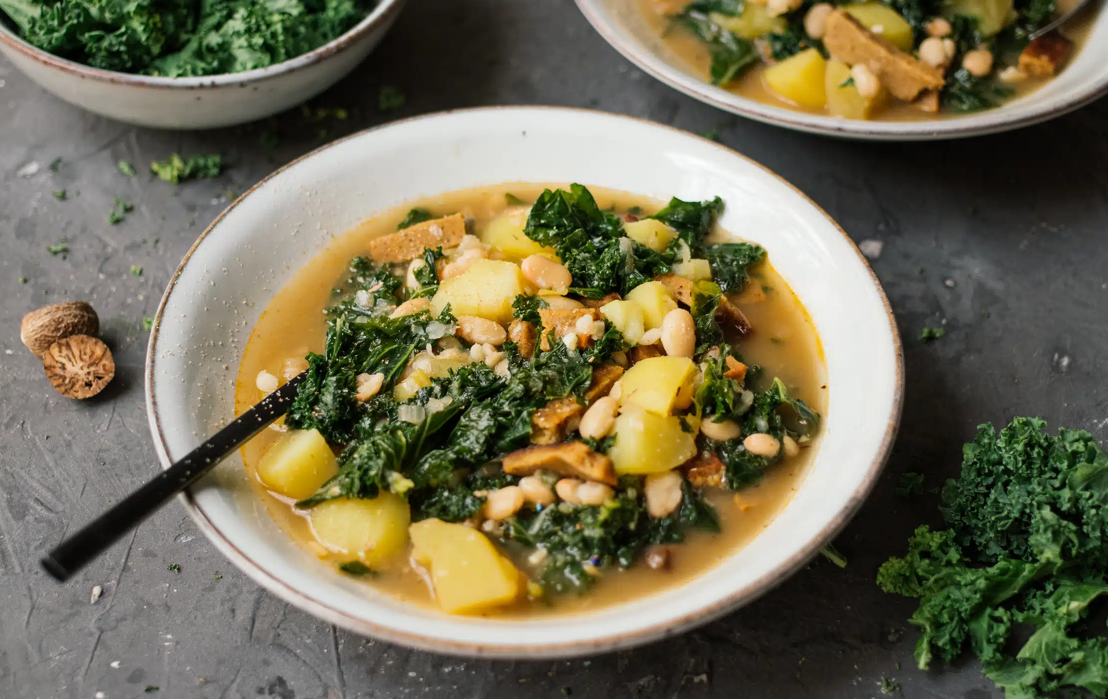 Das Bild zeigt einen Teller mit einem herzhaften **veganen Grünkohleintopf**, der mit Kartoffeln, weißen Bohnen, Pilzen und Grünkohl zubereitet ist. Der Eintopf wirkt cremig und nährstoffreich, garniert mit frischen Kräutern und serviert in einer ansprechenden Schale. Im Hintergrund sind weitere Schüsseln mit Grünkohl zu sehen, was die frische und saisonale Zutatenwahl unterstreicht. Ein perfektes Gericht für eine gesunde, vegane Ernährung!