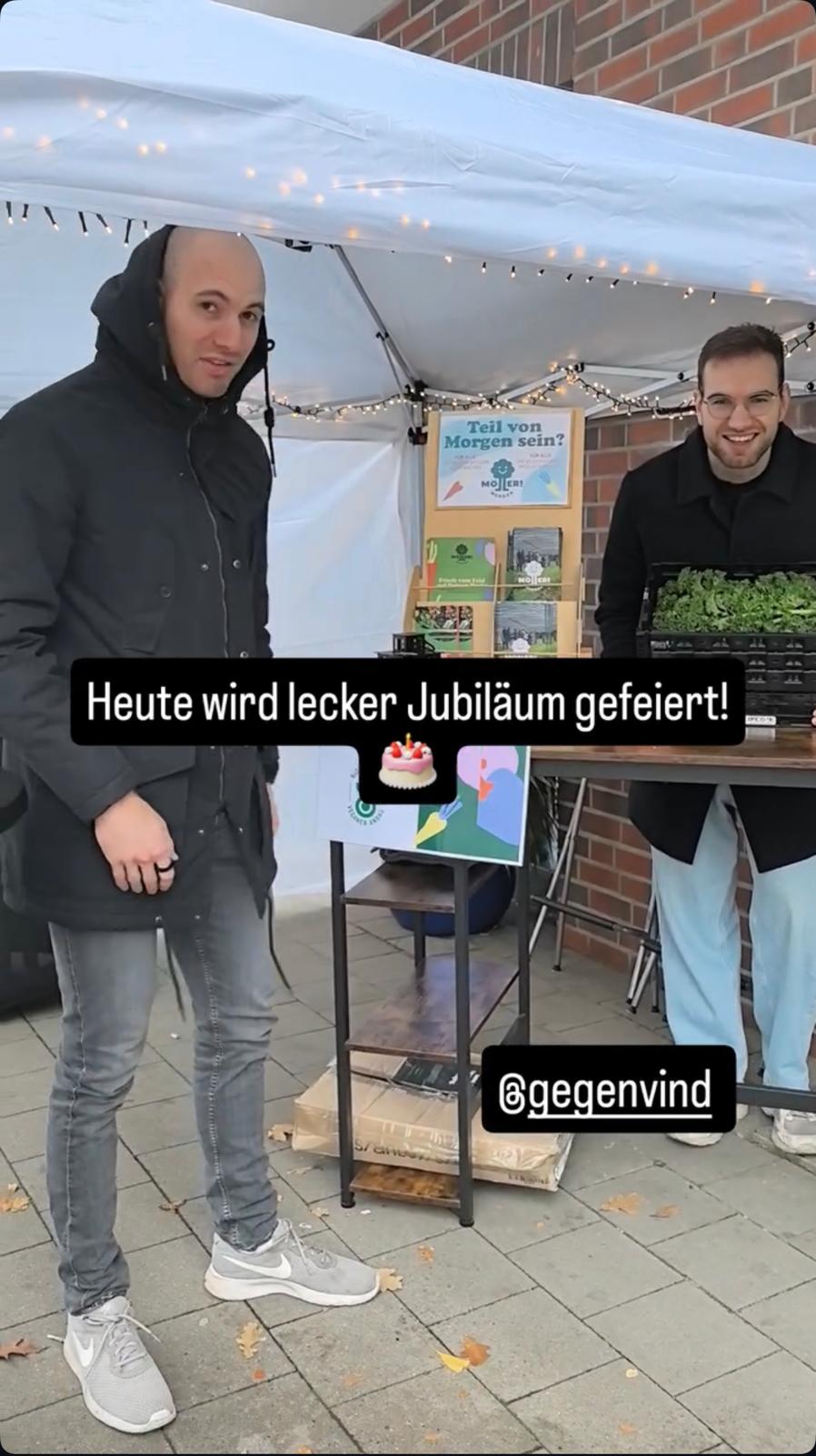 Das Bild zeigt zwei lächelnde Männer an einem Stand, der mit einem Zelt und Lichterketten dekoriert ist. Einer der Männer hält eine Kiste mit frischen Gemüsepflanzen. Im Vordergrund steht der Text:
**"Heute wird lecker Jubiläum gefeiert!"**
Der Stand wirkt einladend und informativ, mit einem Plakat im Hintergrund, das die Frage stellt: **"Teil von Morgen sein?"** sowie dem Logo von **Möllers Morgen**. Zudem ist der Instagram-Account **@gegenvind** eingeblendet.
Die Szene vermittelt den Eindruck eines besonderen Anlasses, bei dem das Jubiläum mit frischen Produkten und einer positiven Atmosphäre gefeiert wird.