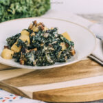 Das Bild zeigt ein leckeres, veganes Gericht aus **Grünkohl mit Kartoffeln und Räuchertofu**. Der Grünkohl ist fein zubereitet und mit Kartoffelstücken sowie geröstetem Räuchertofu gemischt. Das Gericht wirkt herzhaft und nährstoffreich, perfekt für eine gesunde, pflanzliche Ernährung. Die Präsentation auf dem Teller ist ansprechend, und die Kombination aus Grünkohl, Kartoffeln und Räuchertofu bietet eine ausgewogene Mahlzeit. Ein idealer Vorschlag für ein saisonales, veganes Essen!