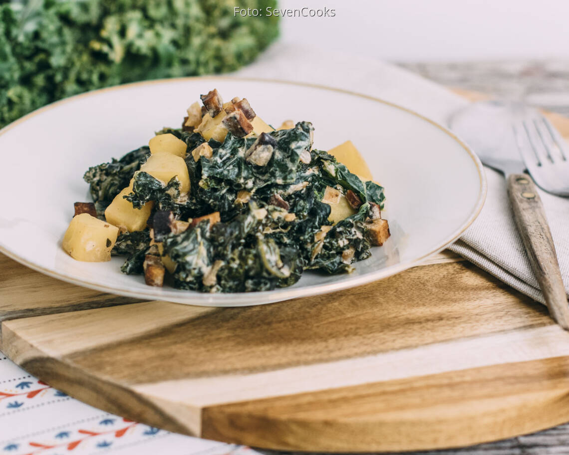 Das Bild zeigt ein leckeres, veganes Gericht aus **Grünkohl mit Kartoffeln und Räuchertofu**. Der Grünkohl ist fein zubereitet und mit Kartoffelstücken sowie geröstetem Räuchertofu gemischt. Das Gericht wirkt herzhaft und nährstoffreich, perfekt für eine gesunde, pflanzliche Ernährung. Die Präsentation auf dem Teller ist ansprechend, und die Kombination aus Grünkohl, Kartoffeln und Räuchertofu bietet eine ausgewogene Mahlzeit. Ein idealer Vorschlag für ein saisonales, veganes Essen!