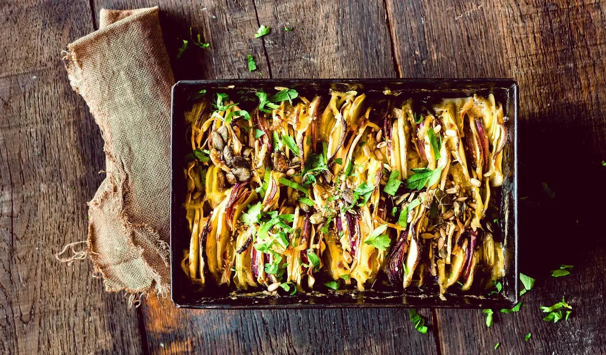 Veganes Kartoffel-Pastinaken-Gratin in einer Backform, überbacken mit goldener Kruste, garniert mit frischen Kräutern und Pilzen, serviert auf einem rustikalen Holztisch mit einem Leinentuch.