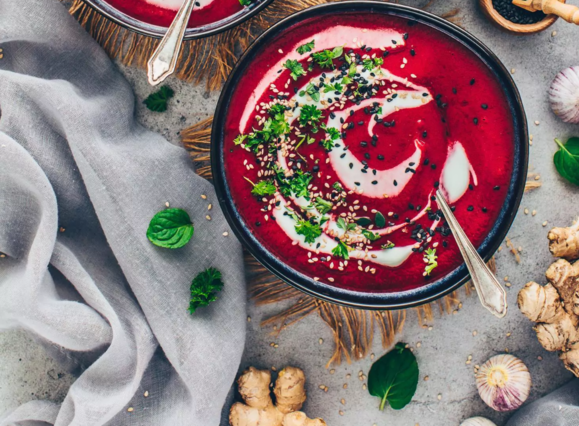 Das Bild zeigt eine wunderschön angerichtete **vegane Rote-Bete-Suppe**. Die Suppe ist cremig, hat eine intensive rote Farbe und ist mit verschiedenen Toppings garniert:
- **Frische Kräuter** wie Petersilie oder Koriander
- **Sesamsamen** und **schwarze Sesamsamen**
- Ein **Schwung Kokosjoghurt oder veganer Sahneersatz**
- **Knoblauchzehen** und **Ingwerstücke** als Dekoration
Die Suppe wird in einer dunklen Schale serviert, die auf einem grauen Untergrund steht. Um die Schale herum sind frische Kräuterblätter, Ingwer und Knoblauchzehen dekorativ platziert.
Diese vegane Rote-Bete-Suppe ist nicht nur optisch ansprechend, sondern auch gesund und voller Geschmack! Möchtest du ein Rezept oder Tipps zur Zubereitung?