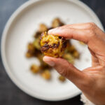 Hand hält ein Stück knusprig gebackenen, smashed Rosenkohl über einem Teller mit weiteren goldbraun gerösteten Rosenkohlstücken.