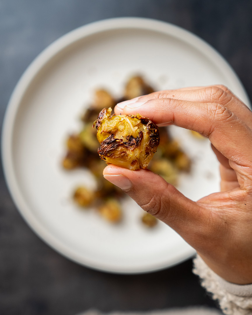 Hand hält ein Stück knusprig gebackenen, smashed Rosenkohl über einem Teller mit weiteren goldbraun gerösteten Rosenkohlstücken.
