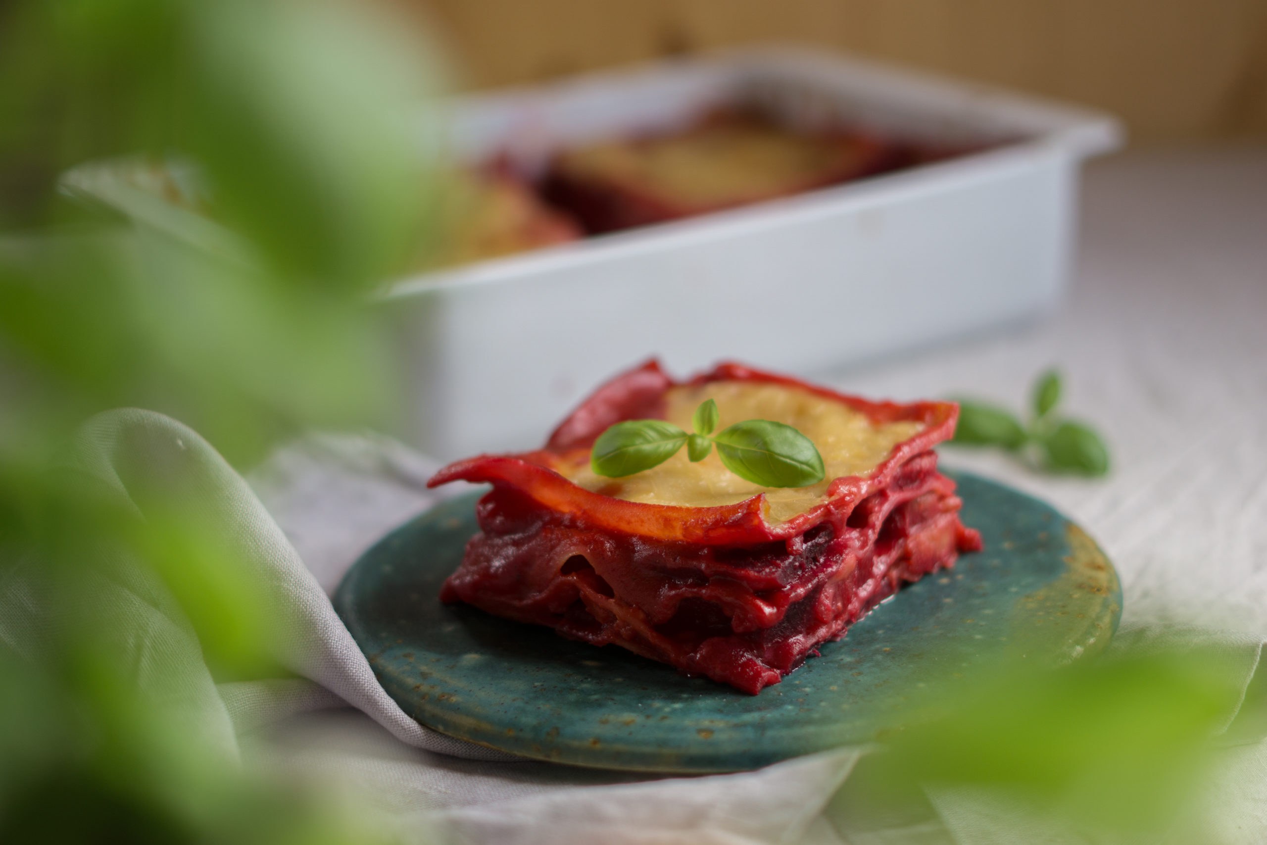 Stück vegane Rote-Bete-Lasagne auf einem grünen Teller, geschichtet mit intensiver Rote-Bete-Füllung und überbacken mit goldener "Käse"-Kruste, garniert mit einem frischen Basilikumblatt. Im Hintergrund steht eine Backform mit der restlichen Lasagne.