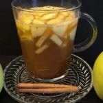 Glas mit warmem Apfel-Zimt-Punsch, gefüllt mit Apfelstücken und Zimtstangen, umgeben von zwei ganzen Äpfeln und einer Zimtstange auf einem dekorativen Untersetzer.