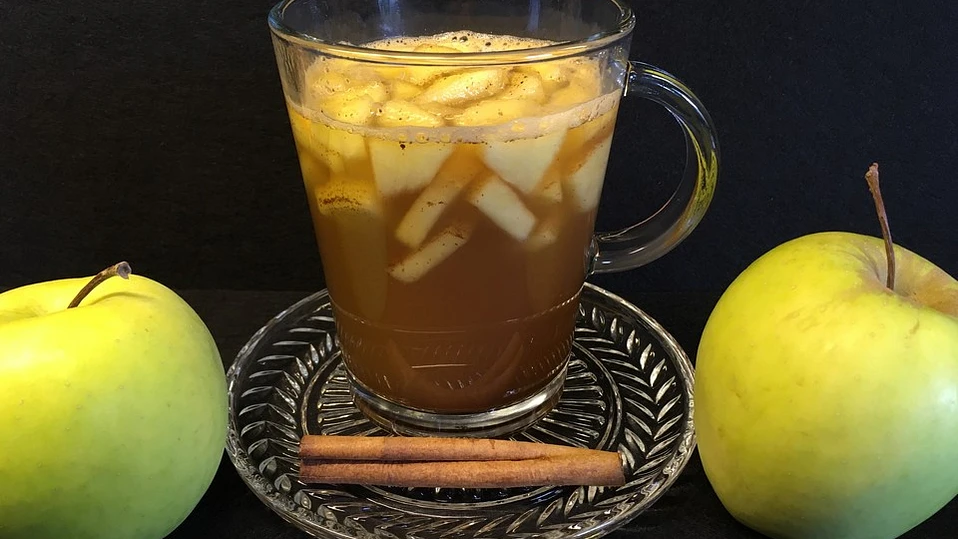 Glas mit warmem Apfel-Zimt-Punsch, gefüllt mit Apfelstücken und Zimtstangen, umgeben von zwei ganzen Äpfeln und einer Zimtstange auf einem dekorativen Untersetzer.