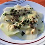 Veganes Kohlrabi-Carpaccio, dünn geschnitten auf einem weißen Teller, garniert mit Sonnenblumenkernen, frischen Kräutern und einer cremigen Sauce.